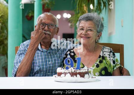 Ältere Paare mit einem Kuchen auf dem Tisch und die Nummer 91 auf dem Kuchen, und der Mann mit einem Finger nach oben Stockfoto