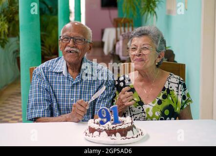 Lächelnde ältere Paare mit einem Kuchen auf dem Tisch und die Nummer 91 auf dem Kuchen Stockfoto