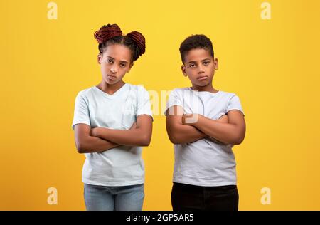 Verärgerte schwarze Jungen und Mädchen mit gekreuzten Armen auf Brust und Blick auf Kamera auf gelb, afroamerikanische Kinder Bruder und Schwester, die negative Emoti ausdrücken Stockfoto