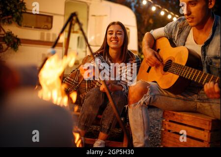 Freunde, die am Lagerfeuer auf der Gitarre spielen, Picknick beim Camping im Wald. Jugend mit Sommerabenteuer auf rv, Camping-Auto auf dem Hintergrund. Zwei Paare Stockfoto