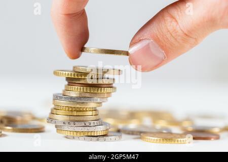Finger, die einen Euro aus nächster Nähe auf den Haufen Münzen legen. Konzept, Geld für die Zukunft zu sparen. Hinzufügen von Bargeld auf Stapel mit weißem Hintergrund. Stockfoto