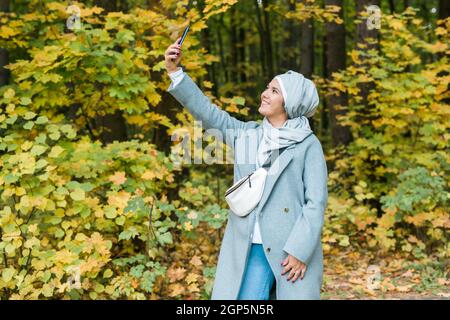 Modische junge muslimische asiatische Mädchen in Hijab nimmt ein Selfie auf dem Smartphone im Freien im Herbst Park Stockfoto