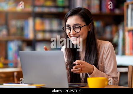 Positive arabische Frau, die eine Videokonferenz auf einem Laptop veranstalte, im Stadtcafe saß, sprach und gestikulte vor der Computer-Webcam. Online-Nachhilfe und Remote-d Stockfoto