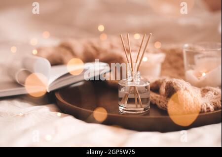 Home Parfüm in Glasflasche mit Holzstäbchen, Duftbrennkerzen, öffnen Sie Papierbuch und stricken Wolltextilien auf Tablett im Schlafzimmer close up. Aromatherapie c Stockfoto