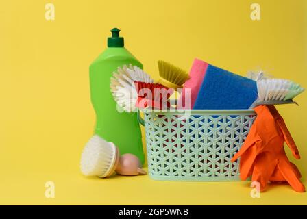 Blauer Kunststoffkorb mit Bürsten, Schwämmen und Gummihandschuhen zum Reinigen, gelber Hintergrund Stockfoto