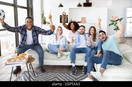 Begeisterte Freunde, die Fußballspiele im Fernsehen sahen, Bierflaschen und Ball hielten und zu Hause Snacks aßen. Gruppe von Sportfans genießen TV-Gam Stockfoto