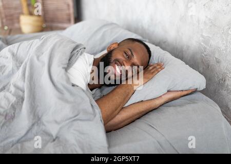 Guten Morgen. Fröhlicher junger afrikanischer Mann, der sich nach dem Aufwachen gut und ausgeruht fühlt, glücklicher bärtiger schwarzer Kerl, der auf der Seite liegt und sich an grauem lin erfreut Stockfoto