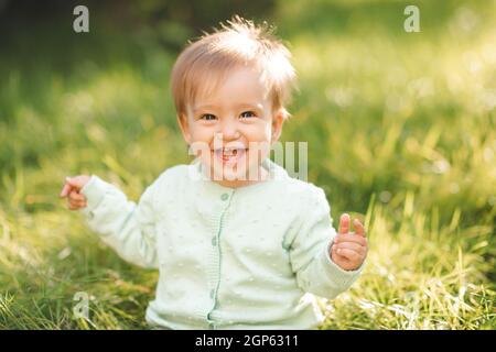 Nette lachende blonde Baby Mädchen 1 Jahre alt tragen Strick Pullover top mit Spaß sitzen im grünen Gras im Freien. Kindheit. Sommersaison. Stockfoto