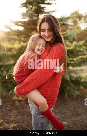 Glückliche Frau halten kleine Kind Mädchen 3-4 Jahre alt tragen rote helle Pullover posiert über Natur Hintergrund aus der Nähe. Lifestyle-Konzept für die ganze Familie. Mutterschaft. A Stockfoto