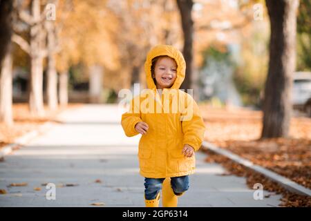 Ziemlich lustig Kind Mädchen 2-3 Jahre alt tragen gelben hellen Regenmantel, Gummistiefel gehen im Park über gefallene Blätter im Freien. Herbstsaison. Frohes Kind über Stockfoto