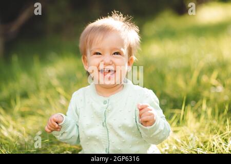 Nette lachende blonde Baby Mädchen 1 Jahre alt tragen Strick Pullover top mit Spaß sitzen im grünen Gras im Freien. Kindheit. Sommersaison. Stockfoto