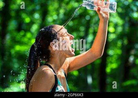 Nahaufnahme einer Läuferin, die nach dem Training Wasser ins Gesicht gießt. Kaltes Wasser aus der Flasche spritzt auf Mädchen Gesicht vor grünem Hintergrund. Stockfoto