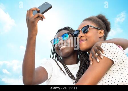 Nahaufnahme Porträt von zwei verschiedenen afrikanischen Teenager-Mädchen tragen Sonnenbrillen nehmen Selbstporträt mit Telefon gegen blauen Himmel. Stockfoto