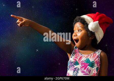 Close up konzeptuelle Porträt von niedlichen afrikanischen Mädchen zeigt auf stars.excited Kind mit offenem Mund trägt weihnachtshut vor dunklen Stern Hintergrund. Stockfoto