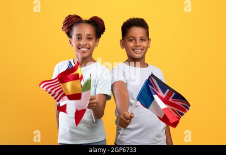 Glückliche schwarze Kinder halten Flaggen verschiedener Länder und posieren zusammen auf gelbem Studiohintergrund. Fröhlicher afroamerikanischer Bruder und Schwester lernten Stockfoto