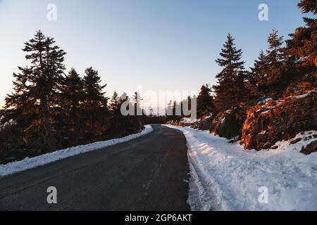 Leere Winter Asphaltstraße bei Sonnenuntergang in der Mitte der bergigen Gegend und Nadelwald schneebedeckt. Hochland von Griechenland, Winterzeit. Freiheit, t Stockfoto