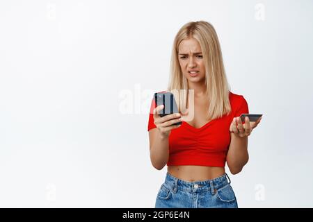 Verwirrte junge Frau, die auf das Mobiltelefon schaut, während sie die Kreditkarte hält, Probleme mit der Bestellung oder Bezahlung hat und auf weißem Hintergrund steht Stockfoto