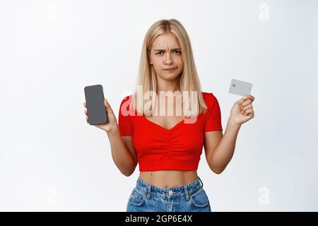 Unzufrieden blonde Mädchen grimaces, zeigt Handy-Bildschirm und Kreditkarte, pouting aufgeregt, stehend über weißem Hintergrund Stockfoto