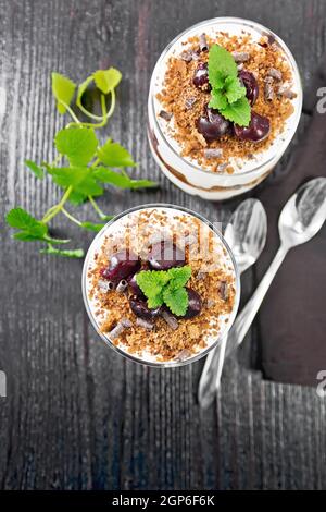 Dessert Schwarzwald von Kirschen, Schokoladenkeks und weichen Quark mit Sahne in zwei Gläsern, Serviette, Minze und Löffel auf Holzbrett Backg Stockfoto