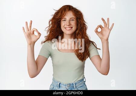 Junge glückliche Frau mit roten langen Haaren, zeigt okay, OK-Zeichen und sieht zufrieden aus, nickt in Zustimmung, macht ja Geste, stehend in T-Shirt über weiß Stockfoto