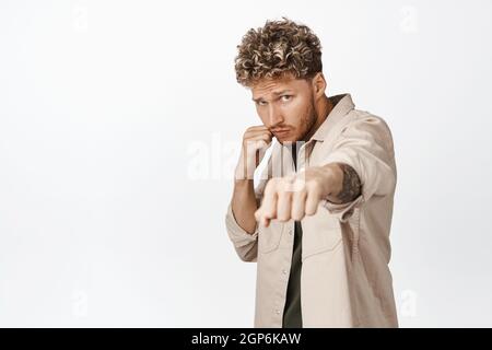 Frech schöner Mann stanzt, knallt Faust und Schatten Boxen, kämpft mit jemandem in legerer Kleidung, stehend über weißem Hintergrund Stockfoto