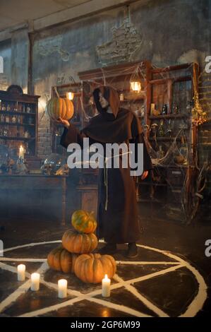 Männlicher Exorzist in schwarzer Kapuze hält Kürbis in der Nähe des magischen Kreises. Exorzismus, Mystery Paranormal Ritual, dunkle Religion, Nacht Horror, Tränke im Regal auf Stockfoto