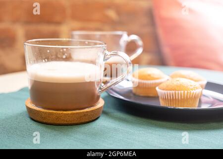 Eine Glastasse heißen Kakao oder Cappuccino auf einem Korkständer, drei Muffins auf einem Teller, stehend auf einem Tisch, beleuchtet durch Licht aus einem Fenster. Frühstück Stockfoto
