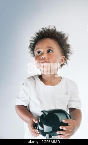 Porträt eines niedlichen kleinen afroamerikanischen Jungen Modell mit Piggy Bank in den Händen. Isoliert auf grauweißem Hintergrund. Investition in die Zukunft. Savi Stockfoto