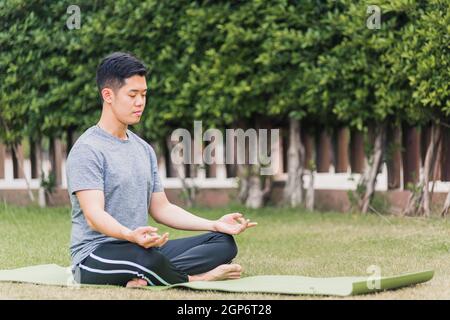 Asiatische junge Mann tun Yoga im Freien in meditieren Lotus Pose sitzen auf grünem Gras mit geschlossenen Augen im Garten Park, Gesundheitskonzept Stockfoto