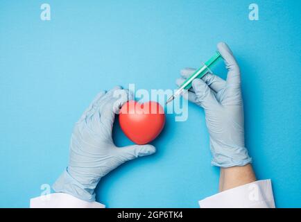 Zwei Hände in blauen Latexhandschuhen halten rotes Herz und eingeklemmte Nadel einer Plastikspritze innen davon auf einem blauen Hintergrund, Draufsicht Stockfoto