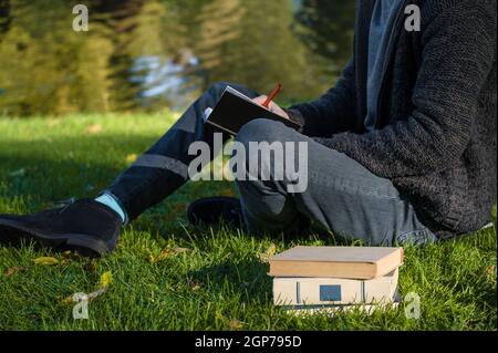 Der Student sitzt im Park auf dem Gras und macht sich Notizen. Ein Stapel Bücher. Teich im Hintergrund. Stockfoto