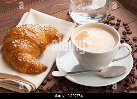 Cappuccino mit Croissant und ein Glas Wasser in die Schale mit dunkelbraunem Holz. Stockfoto