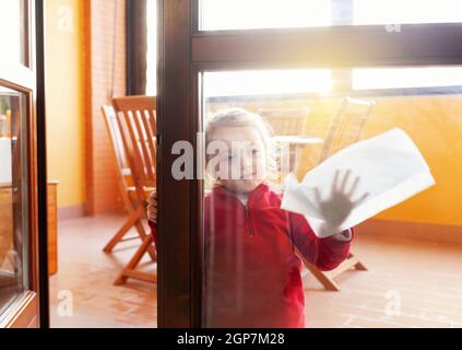 Drei Jahre altes Mädchen hilft bei der Haushaltsreinigung. Sie reinigt die Fenster mit einem Blatt Papier. Idee, die Zeit während des Covid-19 Coronavirus zu vergehen Stockfoto