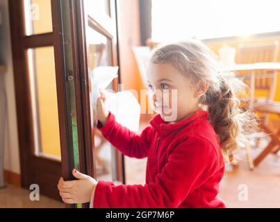 Drei Jahre altes Mädchen hilft bei der Haushaltsreinigung. Sie reinigt die Fenster mit einem Blatt Papier. Idee, die Zeit während des Covid-19 Coronavirus zu vergehen Stockfoto