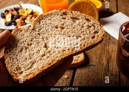 Stück Bio-Vollkornbrot auf rustikalem Tisch. Im Hintergrund getrocknete Früchte, Orangensaft und streichfähige Schokolade. Stockfoto