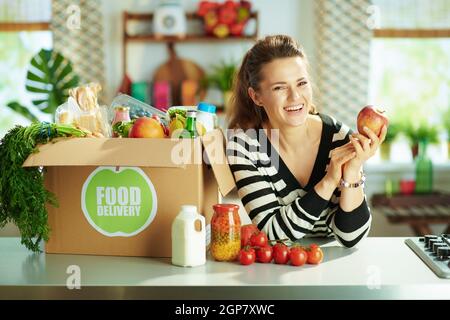 Lieferung von Lebensmitteln. Glückliche moderne 40 Jahre alte Frau mit Lebensmittelbox und Apfel in der Küche. Stockfoto