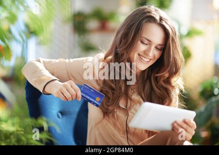 Green Home. Glückliche moderne Frau mit langen welligen Haaren mit Kreditkarte mit Tablet-PC im modernen Wohnzimmer in sonnigen Tag. Stockfoto