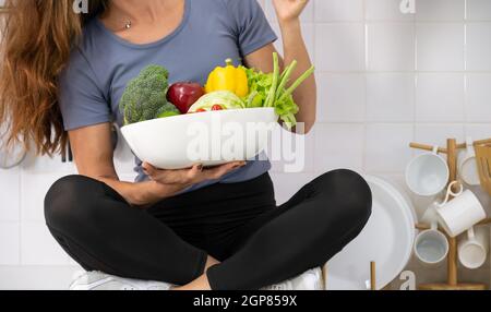 Junge Frau, die mit frischem Gemüse auf der Küchenschranktheke sitzt. Frau in Sportkleidung mit einer weißen Schale mit frischem Gemüse in der Hand. Gesund Stockfoto