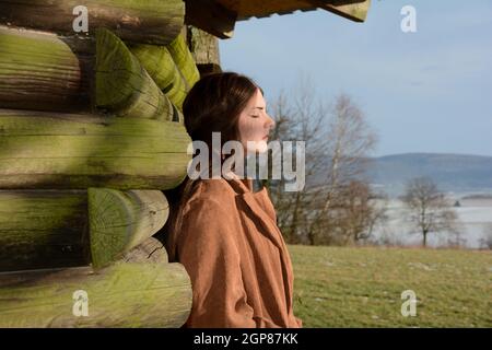 Junge Frau von der Seite, mit hellbraunem Mantel, lehnt an einer Holzhütte, mit einem ernsten Ausdruck auf ihrem Gesicht und geschlossenen Augen, genießen die su Stockfoto