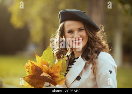 Hallo oktober. Porträt einer glücklichen trendigen Frau im beigen Trenchcoat mit herbstlichen gelben Blättern draußen in der Stadt im Herbst. Stockfoto