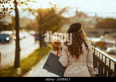 Hallo november. Von hinten gesehen trendige Frau in beigem Trenchcoat und schwarzer Baskenmütze mit Einkaufstaschen und herbstgelben Blättern im Freien auf der City Street Stockfoto