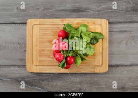 Table Top Aussicht, Bündel von frischen Nass Rettich (raphanus) mit grünen Blättern, Wassertropfen, auf Schneidebrett auf grau Holz Schreibtisch platziert. Stockfoto