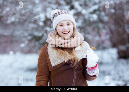 Happy stylish Mädchen in einem gestrickten Hut und Schaffell Mantel mit Fäustlingen und Schneemann im Freien im Stadtpark im Winter. Stockfoto