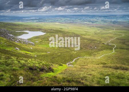 Blick vom Berggipfel auf gewundenen Holzweg des Cuilcagh Park Promenade im Tal unten mit kleinem See, Nordirland Stockfoto