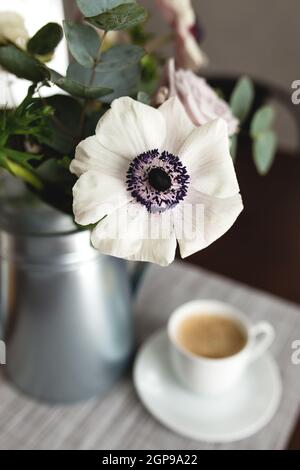 Zarte Anemonen Blumen in einer Metalltopf Vase mit einer Tasse Kaffee auf dem Holztisch. Unscharfer Hintergrund Stockfoto