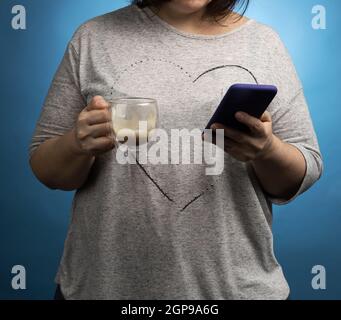 Frau in einem grauen T-Shirt hält eine unvollendete transparente Tasse mit Kaffee und einem Smartphone, blauer Hintergrund Stockfoto