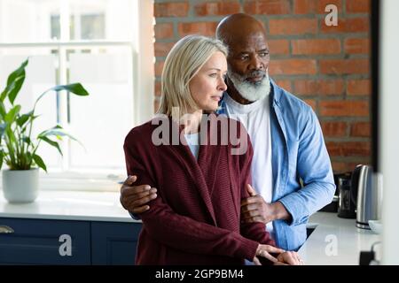Besorgt Senior diverse Paar in der Küche lehnte sich auf Arbeitsplatte, umarmt und wegschaut Stockfoto