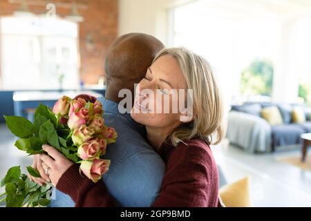 Glückliches, vielseitiges Paar im Wohnzimmer, umarmt und hält Blumen Stockfoto