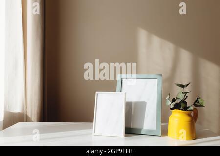 Ein gelber Keramikkrug oder eine Vase mit Eukalyptuszweigen, leere weiße Bilderrahmen auf dem weißen Tisch im Innenraum mit beigen Wänden in der Nähe des Fensters. Stockfoto