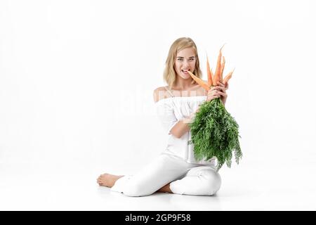 Schöne blonde Frau in einer weißen Bluse hält eine frische Karotte mit grünen Blättern auf weißem Hintergrund. Gesundheit und Ernährung Stockfoto